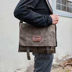 A person in a black jacket and gray pants is holding a brown GORUCK Heritage Claymore Sling Bag, demonstrating its timeless durability. The bag has two straps and is worn over the shoulder. The background features a white wall and a window.