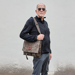An older man in sunglasses and a black jacket stands against a gray wall, holding the GORUCK Heritage Claymore Sling Bag known for its durable craftsmanship.