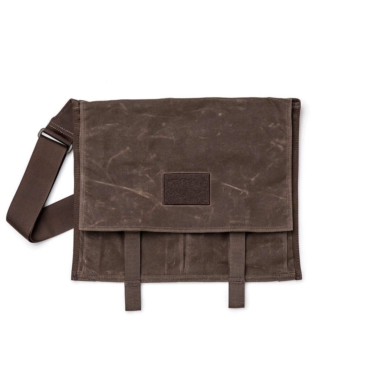 The GORUCK Heritage Claymore Sling Bag boasts a classic brown waxed canvas exterior with an adjustable strap and stitched detailing on the flap. At the center, there's a rectangular leather patch bordered by two strap loops at the bottom.