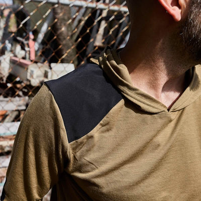 A person wearing the GORUCK Men's Commando Pullover - Merino Wool, featuring a tan and black design with reinforced paneling, stands in front of a chain-link fence. The sun casts shadows over their attire and the fence, highlighting their upper body as they turn their head slightly to the side.