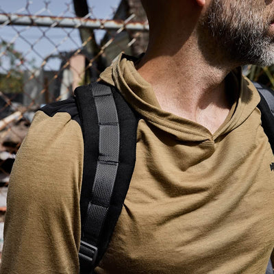 A bearded man in a tan GORUCK Men’s Commando Pullover - Merino Wool and carrying a black backpack stands before a chain-link fence. The focus is on his upper body, highlighting the textures of his clothing, which suggests durability amidst the outdoor setting.