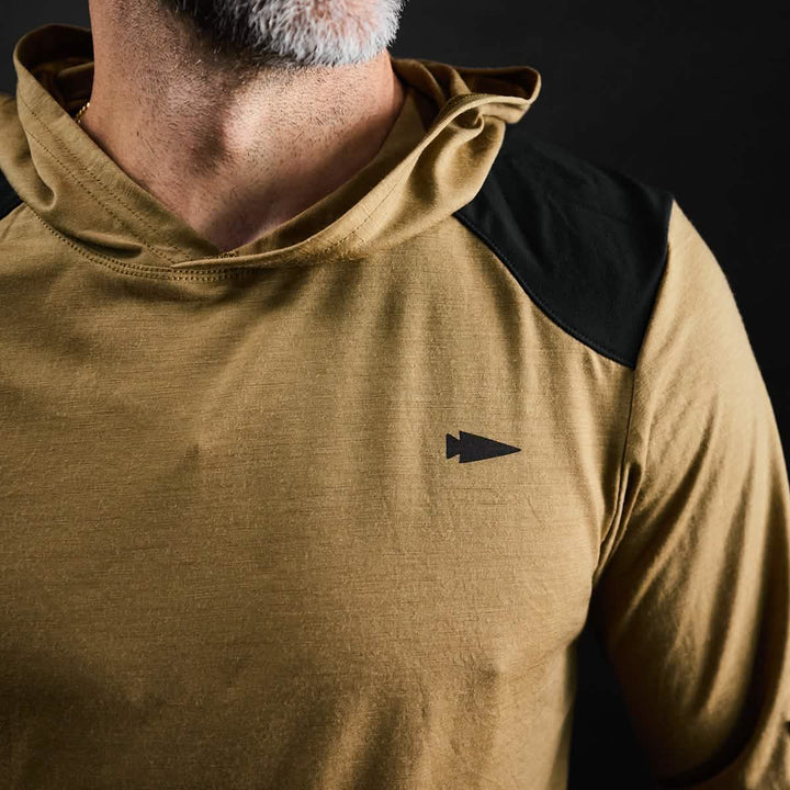 A person is dressed in the "Men’s Commando Pullover - Merino Wool" by GORUCK, featuring a tan hue with black shoulder accents and reinforced paneling. The pullover is adorned with a small black arrowhead logo on the chest. Set against a dark background, the focus is drawn to the apparel, while only part of the person's head can be seen.