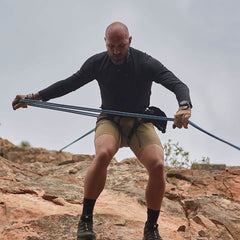 A man is rock climbing, wearing a black long-sleeve shirt and Men’s Simple Shorts from GORUCK in fast-drying beige. He grips the ropes while descending a rocky surface on a cloudy day. With focus in his eyes and equipped with climbing gear, he benefits from the Lightweight ToughDry® fabric in his attire.