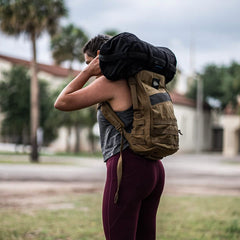 A person with long hair carries a black duffel bag made from ToughMesh™ fabric on their shoulder and wears a brown backpack. Outdoors on a cloudy day with blurred trees and buildings in the background, they are dressed in a featherweight training shirt alongside maroon leggings, complementing their ensemble with the Women's USA Performance Tank by GORUCK.