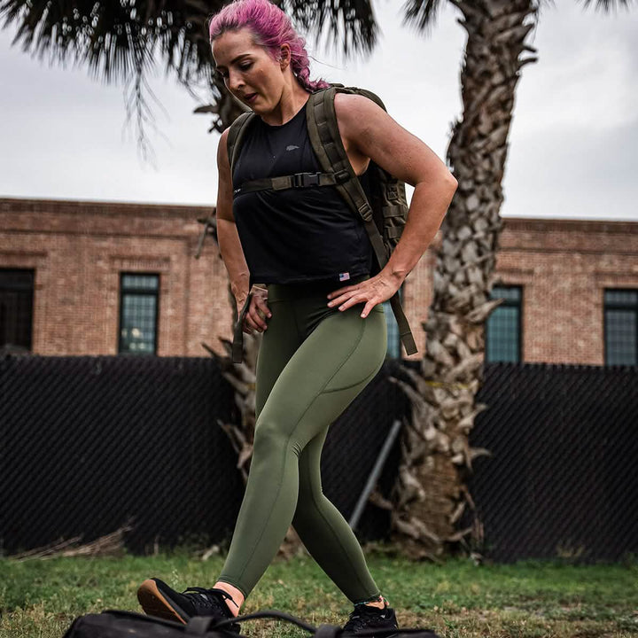 A person with pink hair is wearing a Women’s USA Performance Tank - ToughMesh from GORUCK, green leggings, and a backpack while exercising outdoors. They are stepping purposefully on the grass near tall palm trees and a brick building.