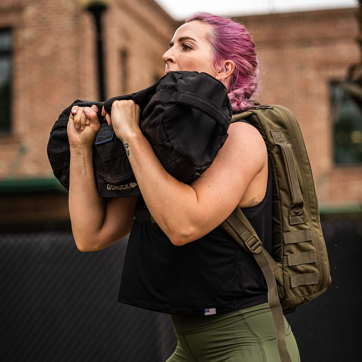 Effortlessly carrying a large black sandbag over their shoulder, a person with pink hair sports a green backpack outdoors in front of a brick building. Clad in green pants and the Women's USA Performance Tank by GORUCK featuring ToughMesh™ fabric, they conquer the day with style and ease.