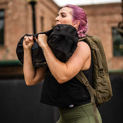 Effortlessly carrying a large black sandbag over their shoulder, a person with pink hair sports a green backpack outdoors in front of a brick building. Clad in green pants and the Women's USA Performance Tank by GORUCK featuring ToughMesh™ fabric, they conquer the day with style and ease.