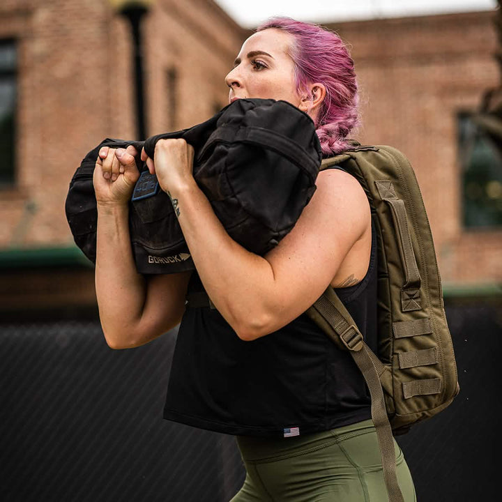 Effortlessly carrying a large black sandbag over their shoulder, a person with pink hair sports a green backpack outdoors in front of a brick building. Clad in green pants and the Women's USA Performance Tank by GORUCK featuring ToughMesh™ fabric, they conquer the day with style and ease.