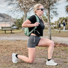 Woman lunging outdoors wearing GORUCK backpack, sunglasses, black top, gray shorts, white sneakers in park