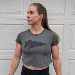 fit woman in grey cropped GORUCK t-shirt with dark arrowhead logo standing against textured garage door