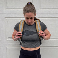 Woman adjusting tan rucksack straps wearing dark gray crop top and black leggings against white garage door