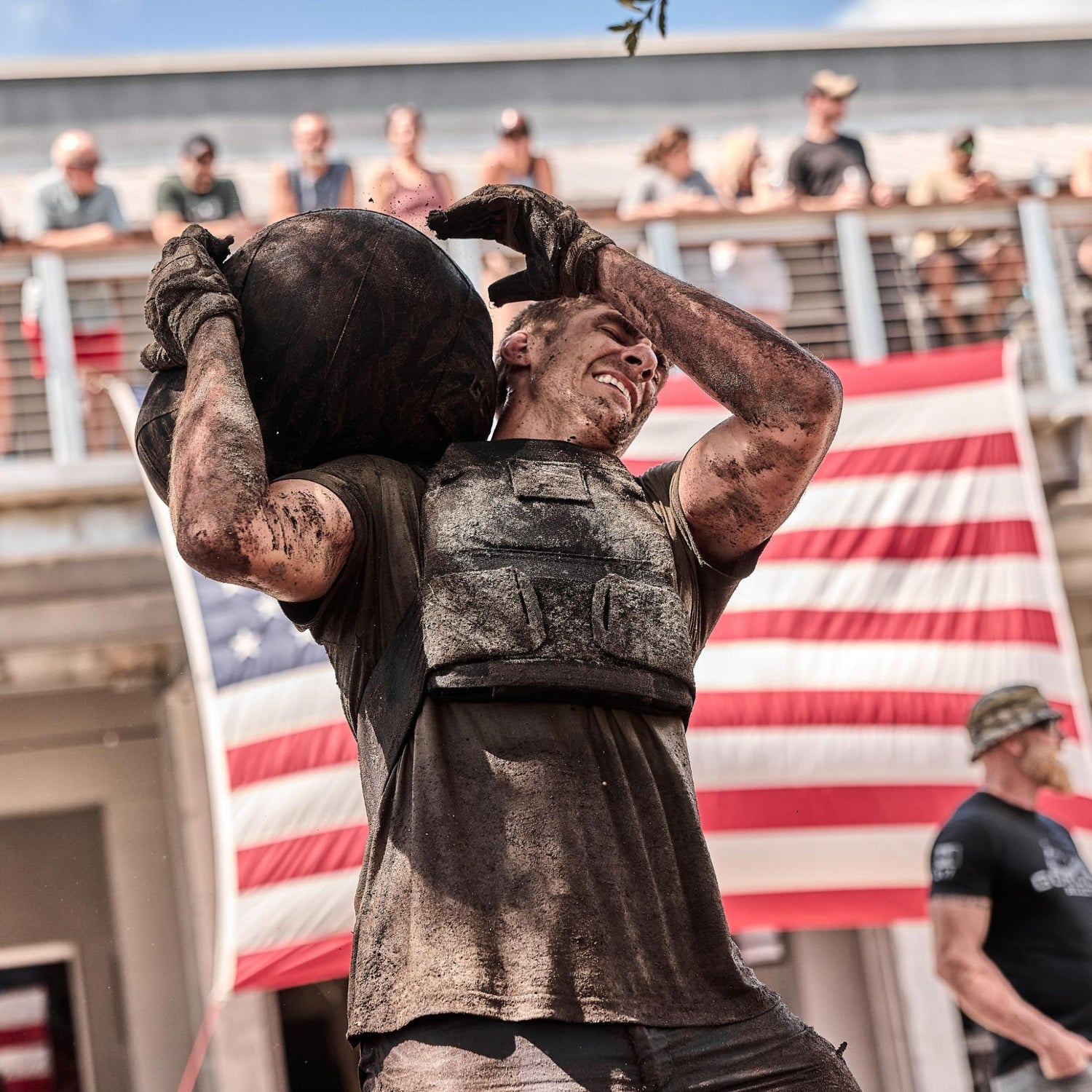 A muddy athlete in a vest lifts a heavy ball overhead, with an American flag in the background.