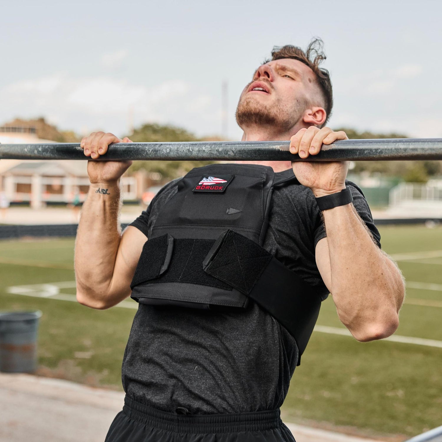 Man wearing a weighted vest doing a pull-up outdoors on a metal bar at a sports field.