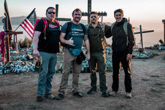 Four men wearing GORUCK rucking gear posing at a memorial site with crosses and an American flag at sunset