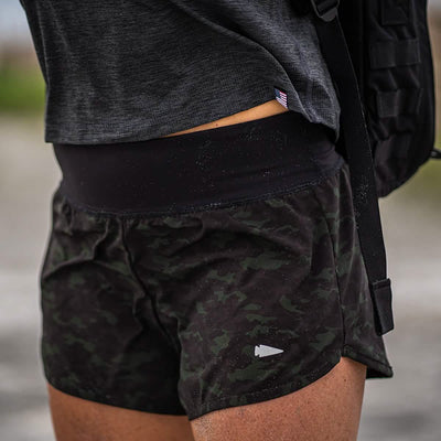 A person dressed in GORUCK's Women’s Training Shorts - ToughStretch in dark camo and a gray shirt is carrying a backpack. The image highlights the shorts, crafted from durable ToughStretch fabric, emphasizing their snug fit and squat-proof design, with a subtle arrow logo clearly visible.