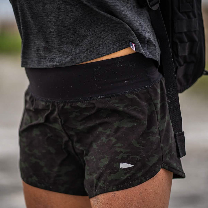 A person dressed in GORUCK's Women’s Training Shorts - ToughStretch in dark camo and a gray shirt is carrying a backpack. The image highlights the shorts, crafted from durable ToughStretch fabric, emphasizing their snug fit and squat-proof design, with a subtle arrow logo clearly visible.