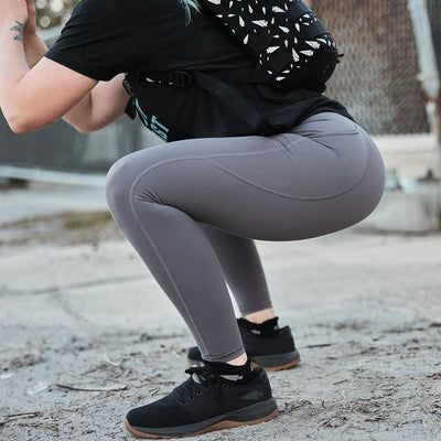 A person in GORUCK's Women’s Training Leggings Pocket - ToughFlex in gray and a black shirt is squatting outdoors, wearing black sneakers. The scene includes a background with a chain-link fence and concrete, while they carry a patterned backpack.
