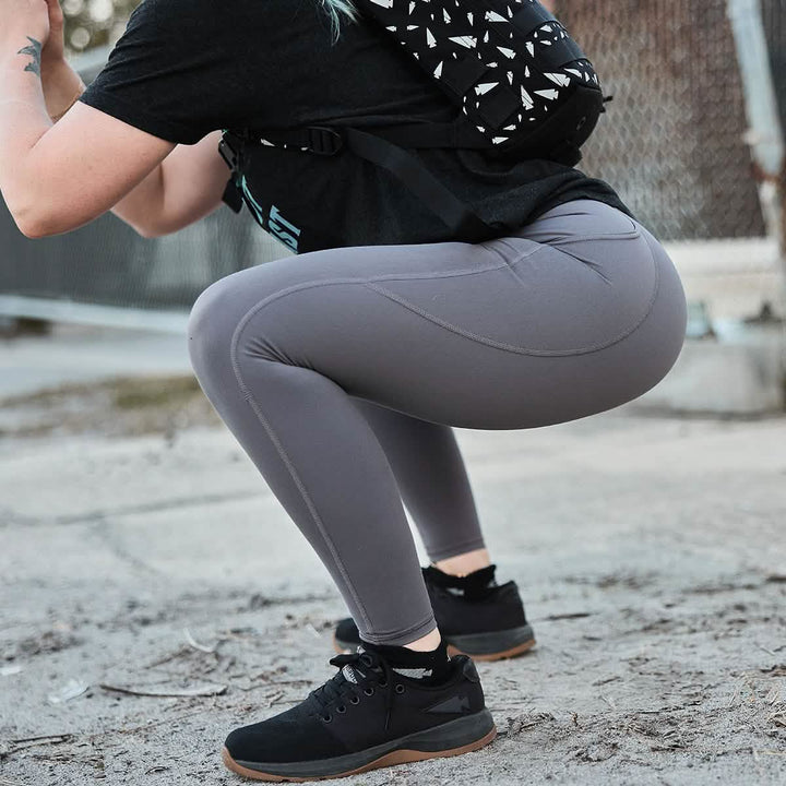 A person in GORUCK's Women’s Training Leggings Pocket - ToughFlex in gray and a black shirt is squatting outdoors, wearing black sneakers. The scene includes a background with a chain-link fence and concrete, while they carry a patterned backpack.