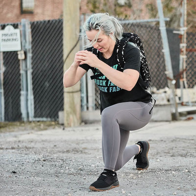 A person with light hair kneels while exercising outside, wearing a black T-shirt, GORUCK Women’s Training Leggings Pocket - ToughFlex, and black sneakers. They have a black backpack, and the background features a chain-link fence and a Private Parking sign.