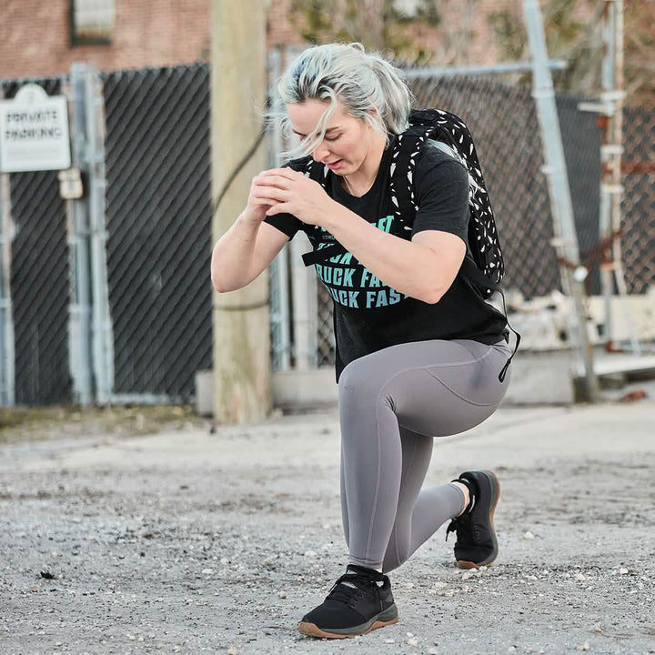 A person with light hair kneels while exercising outside, wearing a black T-shirt, GORUCK Women’s Training Leggings Pocket - ToughFlex, and black sneakers. They have a black backpack, and the background features a chain-link fence and a Private Parking sign.