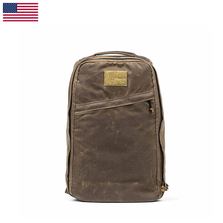 The GR1 USA - Heritage by GORUCK is a brown, rectangular rucksack made from waxed duck canvas. It features multiple zippers, a top handle, and a small patch on the front displaying a U.S. flag in the top left corner against a white background.