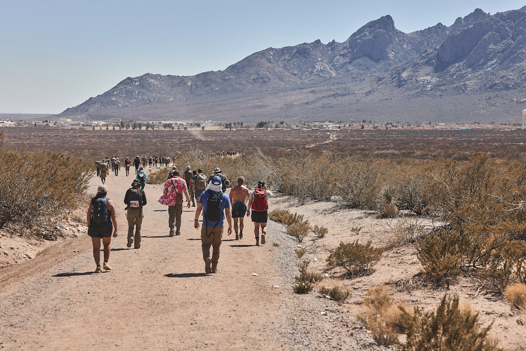 People walking on a dirt path in a desert landscape with mountains in the background under a clear sky.