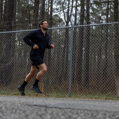 Man running outdoors in black athletic gear near chain-link fence with forest background