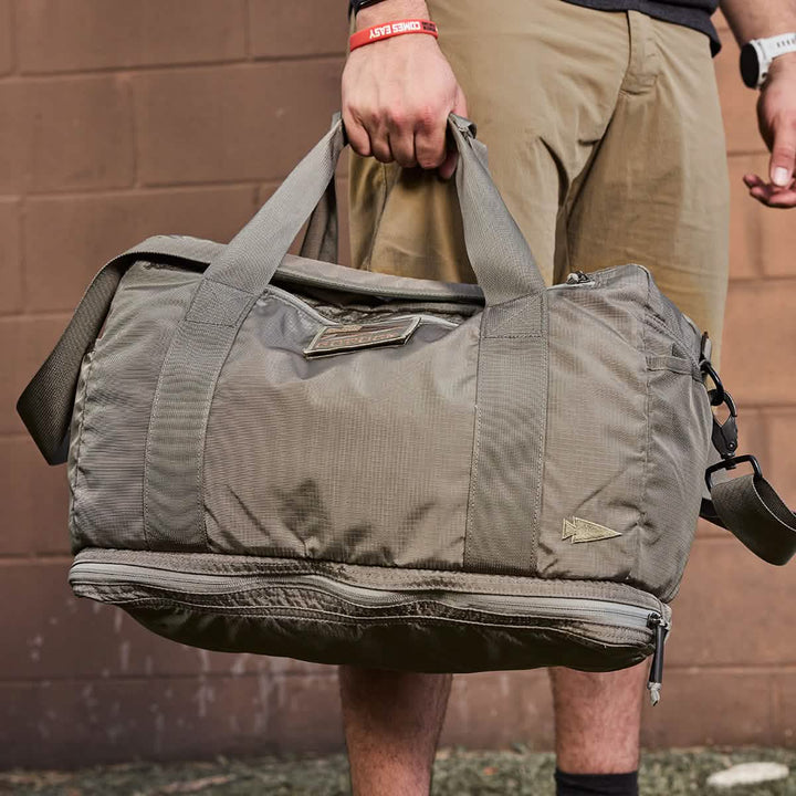 A person holding a green GORUCK Kit Bag w/ Shoe Compartment - Ripstop ROBIC® with handles and a shoulder strap, stands in front of a brown brick wall. The bag features multiple zippers and pockets. The individual is wearing a red wristband, black shirt, beige pants, and a watch.