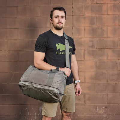 A person stands against a brick wall with a GORUCK Kit Bag with Shoe Compartment - Ripstop ROBIC® slung over their shoulder, complete with a shoulder strap. They wear a black T-shirt that features a fish graphic and the word GOFISH, paired with khaki shorts and a wristwatch.