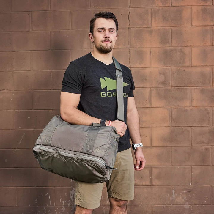 A person stands against a brick wall with a GORUCK Kit Bag with Shoe Compartment - Ripstop ROBIC® slung over their shoulder, complete with a shoulder strap. They wear a black T-shirt that features a fish graphic and the word GOFISH, paired with khaki shorts and a wristwatch.