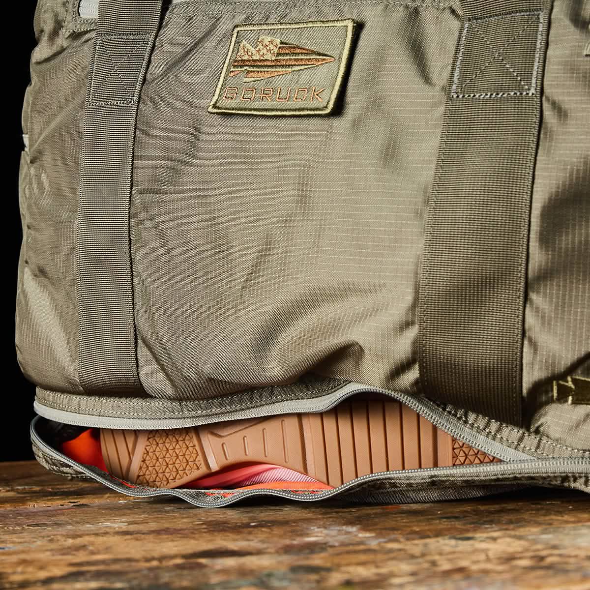 A khaki-colored Kit Bag with a Shoe Compartment, made from Ripstop ROBIC® fabric by GORUCK, features a patch showcasing an airplane image and the word GORUCK. The bag is partially open to reveal the sole of an orange and brown athletic shoe inside. Resting on a wooden surface, it captures the rugged appeal of a Special Forces Flight Bag.