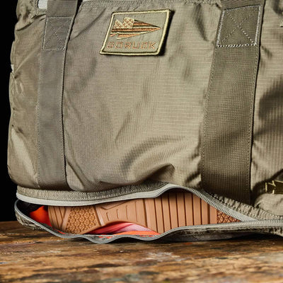 A khaki-colored Kit Bag with a Shoe Compartment, made from Ripstop ROBIC® fabric by GORUCK, features a patch showcasing an airplane image and the word GORUCK. The bag is partially open to reveal the sole of an orange and brown athletic shoe inside. Resting on a wooden surface, it captures the rugged appeal of a Special Forces Flight Bag.