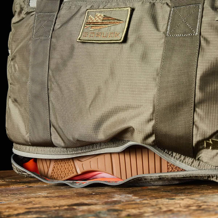 A khaki-colored Kit Bag with a Shoe Compartment, made from Ripstop ROBIC® fabric by GORUCK, features a patch showcasing an airplane image and the word GORUCK. The bag is partially open to reveal the sole of an orange and brown athletic shoe inside. Resting on a wooden surface, it captures the rugged appeal of a Special Forces Flight Bag.