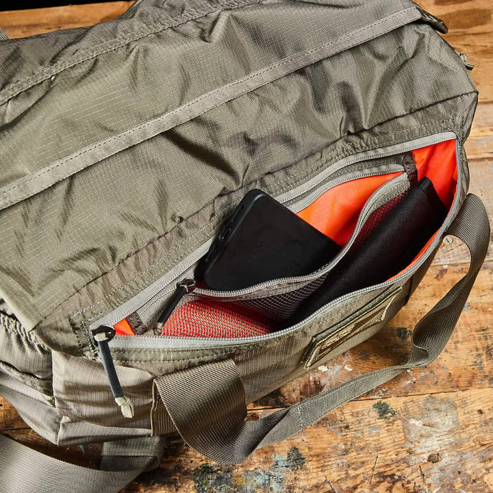A khaki Kit Bag with Shoe Compartment by GORUCK is placed on a wooden surface, its side pocket unzipped to reveal an orange interior lining and a black smartphone.
