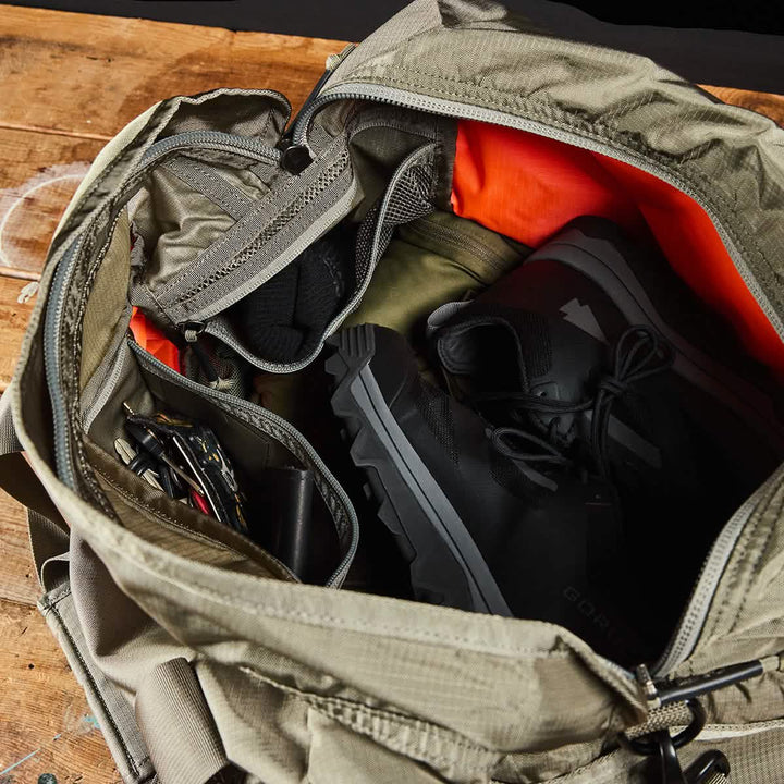 A khaki GORUCK Kit Bag made from Ripstop ROBIC® fabric with an included shoulder strap is open on a wooden surface, showing its orange interior. Inside, there are black hiking boots in the shoe compartment, a rolled-up green jacket, black socks, and various tools stowed in a side pocket.