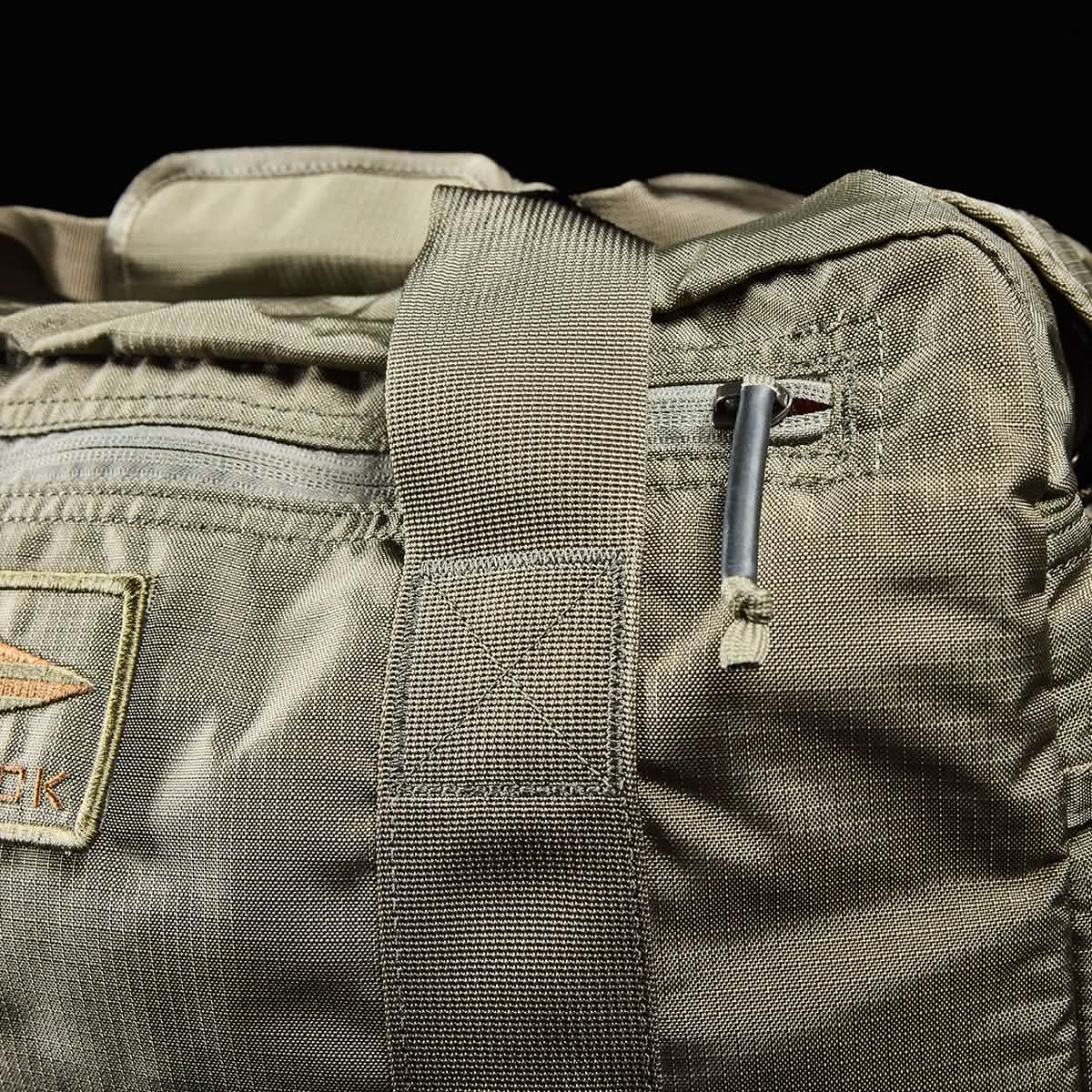 Close-up of a khaki-colored Kit Bag with Shoe Compartment by GORUCK, featuring a durable shoulder strap and a small pocket. A partial view of the stitched logo is visible on the left, while the zipper pull at the top accentuates its rugged design reminiscent of deployment bags.