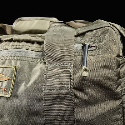Close-up of a khaki-colored Kit Bag with Shoe Compartment by GORUCK, featuring a durable shoulder strap and a small pocket. A partial view of the stitched logo is visible on the left, while the zipper pull at the top accentuates its rugged design reminiscent of deployment bags.