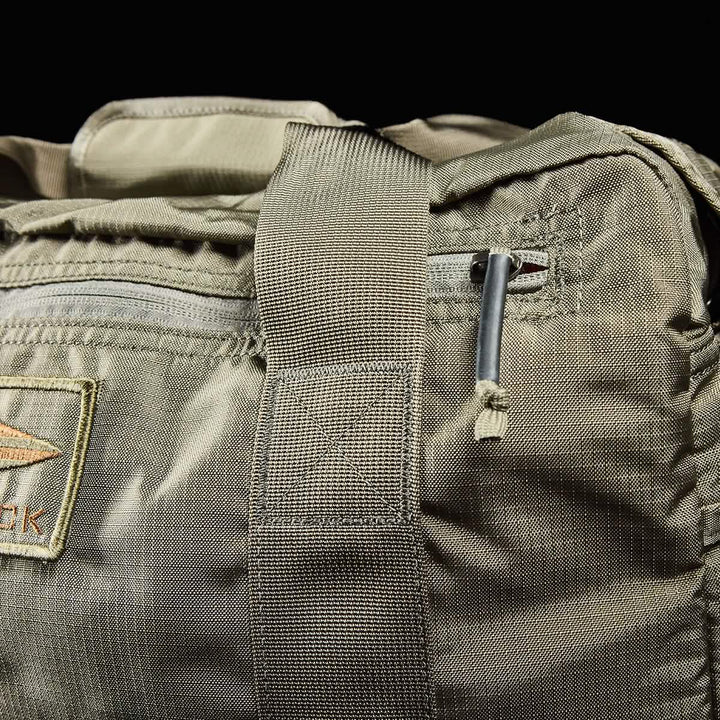 Close-up of a khaki-colored Kit Bag with Shoe Compartment by GORUCK, featuring a durable shoulder strap and a small pocket. A partial view of the stitched logo is visible on the left, while the zipper pull at the top accentuates its rugged design reminiscent of deployment bags.