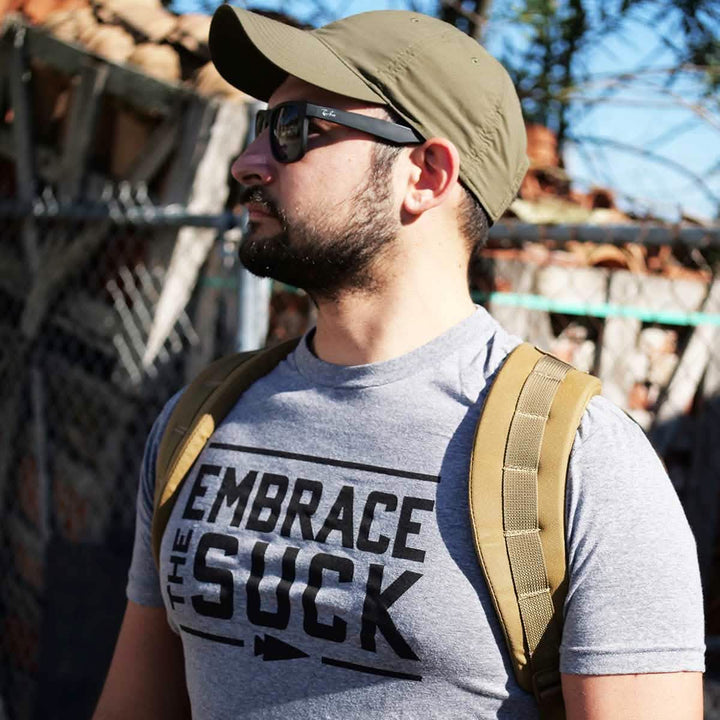 A man confidently stands outside near a chain-link fence, wearing USMC sunglasses, a green cap, and the vendor-unknown Embrace the Suck Tee in tri-blend gray. With a tan backpack slung over his shoulder, he is framed against a sunny sky and brick backdrop.