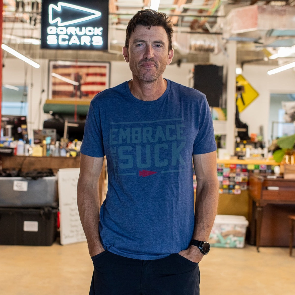 A man wearing the Embrace the Suck Tee - Tri-BlendX in blue stands indoors in front of a GORUCK SCARS sign.
