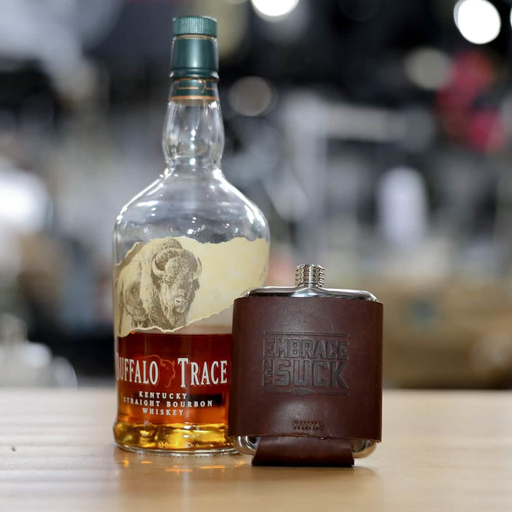 A bottle of Buffalo Trace bourbon, partially filled, stands beside GORUCK's Leather Wrapped Flask – Built By Scars, adorned with "Embrace the Suck" embossed on its leather wrap. Both rest on a wooden surface with a blurred background, suggesting an adventure waiting to unfold.