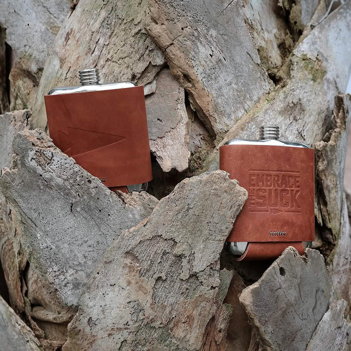 Amidst large pieces of bark, two GORUCK Leather Wrapped Flasks - Built By Scars rest easily, crafted from stainless steel and wrapped in leather. One flask boasts the embossed text EMBRACE THE SUCK, embodying the spirit of adventure and seamlessly blending into the rustic, natural texture around it.