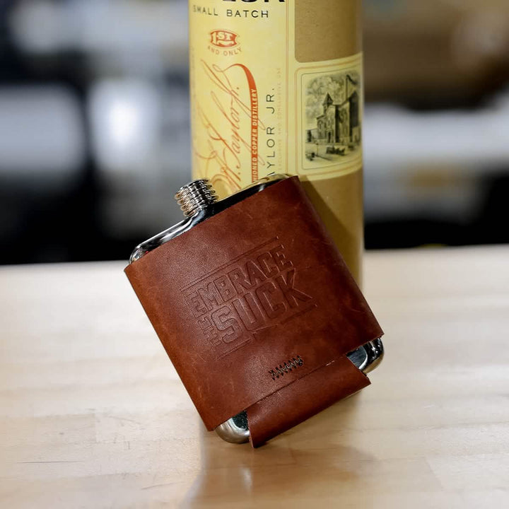 A Leather Wrapped Flask - Built By Scars by GORUCK, made from stainless steel and featuring EMBRACE THE SUCK embossed on its surface, stands in front of a partially visible bottle with an intricate label. Both the flask and the bottle sit on a wooden surface, capturing the essence of adventure and rugged elegance.