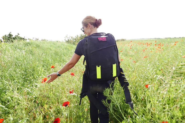 A woman with a black backpack walks through a field of tall grass and red poppies, reaching out to the flowers.