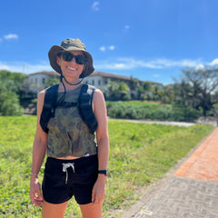 A smiling person wearing the Boonie Hat - Tactical - ToughDry, sunglasses, and a backpack stands outdoors on a sunny day, surrounded by lush greenery.