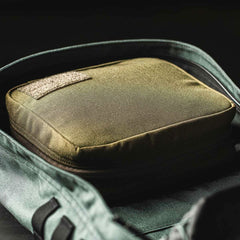 Inside the open backpack lies a green GORUCK GR2 Field Pocket, featuring a textured patch and padded design. The backpack, crafted from 1000D Cordura, is partially visible with its black interior contrasted against a light green exterior. Soft lighting casts gentle shadows that accentuate the fabric's texture.