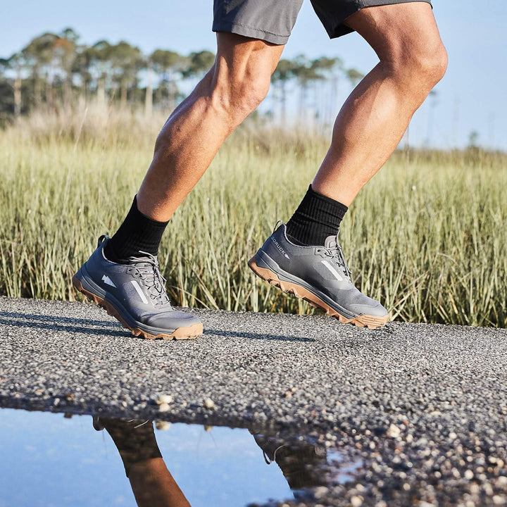 A person in Mackall sneakers by GORUCK, featuring Forged Iron, Chiseled Stone, and Gum hues, ideal for all-terrain adventures, is jogging outdoors. The background reveals tall grass and trees while the gravelly ground showcases a reflective puddle nearby.