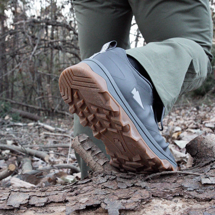 A close-up shows a person hiking through a wooded area, stepping over a fallen log in GORUCK's Mackall shoes in Forged Iron and Chiseled Stone with Gum textured soles. The gray shoes pair well with green pants against a backdrop of vibrant trees and leaves.