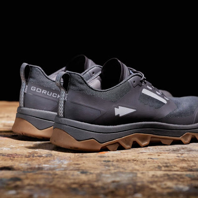 A pair of Mackall trail shoes by GORUCK, in a striking forged iron and chiseled stone color with gum soles, are showcased on a wooden surface. These all-terrain athletic shoes highlight a modern design with branding on the heel and arrow-like graphics along the side, making them ideal for stylishly navigating diverse landscapes.