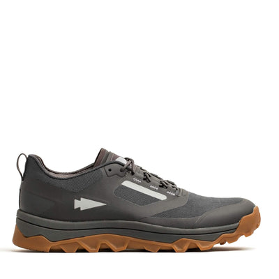 The GORUCK Mackall in Forged Iron, Chiseled Stone, and Gum is a trail shoe crafted for outdoor activities. It boasts a minimalist design with a gray and black color scheme, complemented by a brown sole. The all-terrain textured outsole ensures excellent grip, and a small arrow symbol adorns the side. The background is plain white.